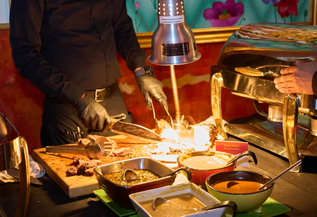 A person wearing black clothes and gloves carves meat at a buffet station, surrounded by various sauces in bowls, under a heat lamp with another person’s hand reaching towards the food.