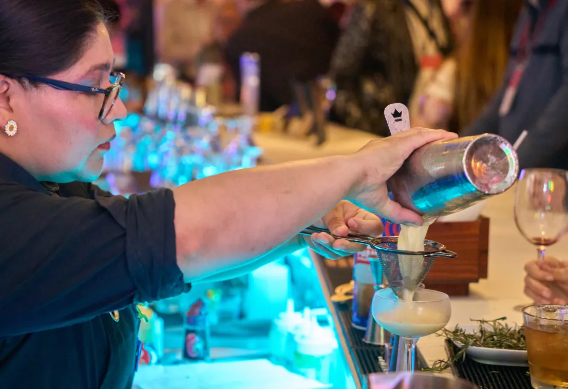 A bartender pours a cocktail through a strainer into a glass at a busy, brightly lit bar while patrons watch from across the counter.