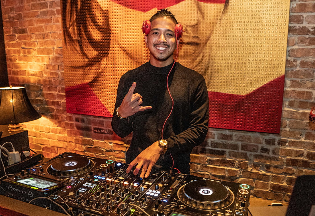 A smiling DJ wearing red headphones and a black sweater stands behind a DJ controller in front of a brick wall, making a hand gesture with one hand while the other hand is on the mixer.