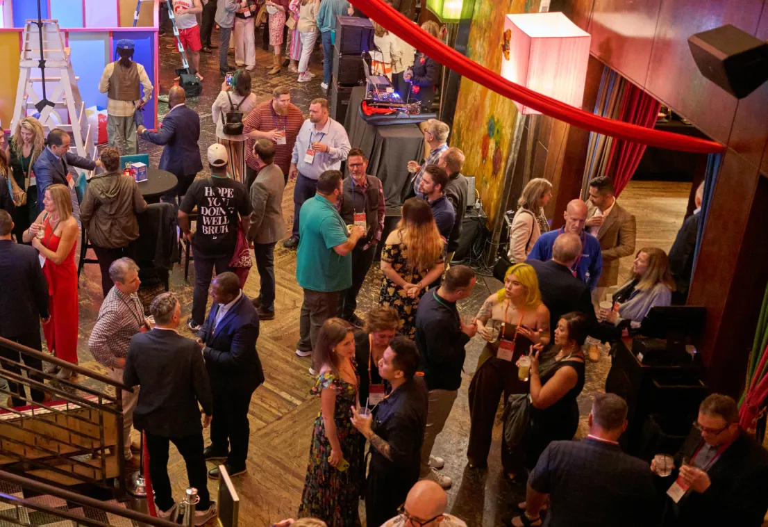 A large group of people socialize and mingle at an indoor event, holding drinks and conversing in a lively, warmly lit room with wooden floors and colorful decor.