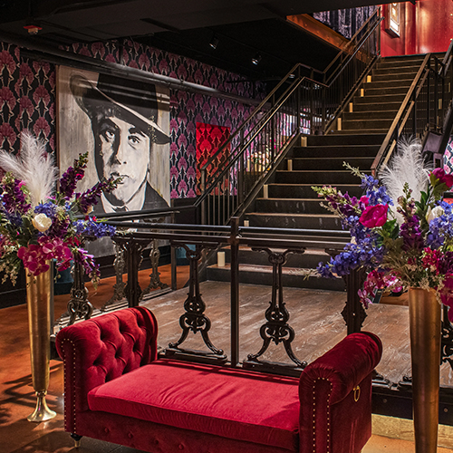 A stylish interior features a red velvet couch, flower arrangements in gold vases, wallpaper with purple designs, and a staircase. A black-and-white portrait of a man wearing a hat hangs on the wall near the stairs.